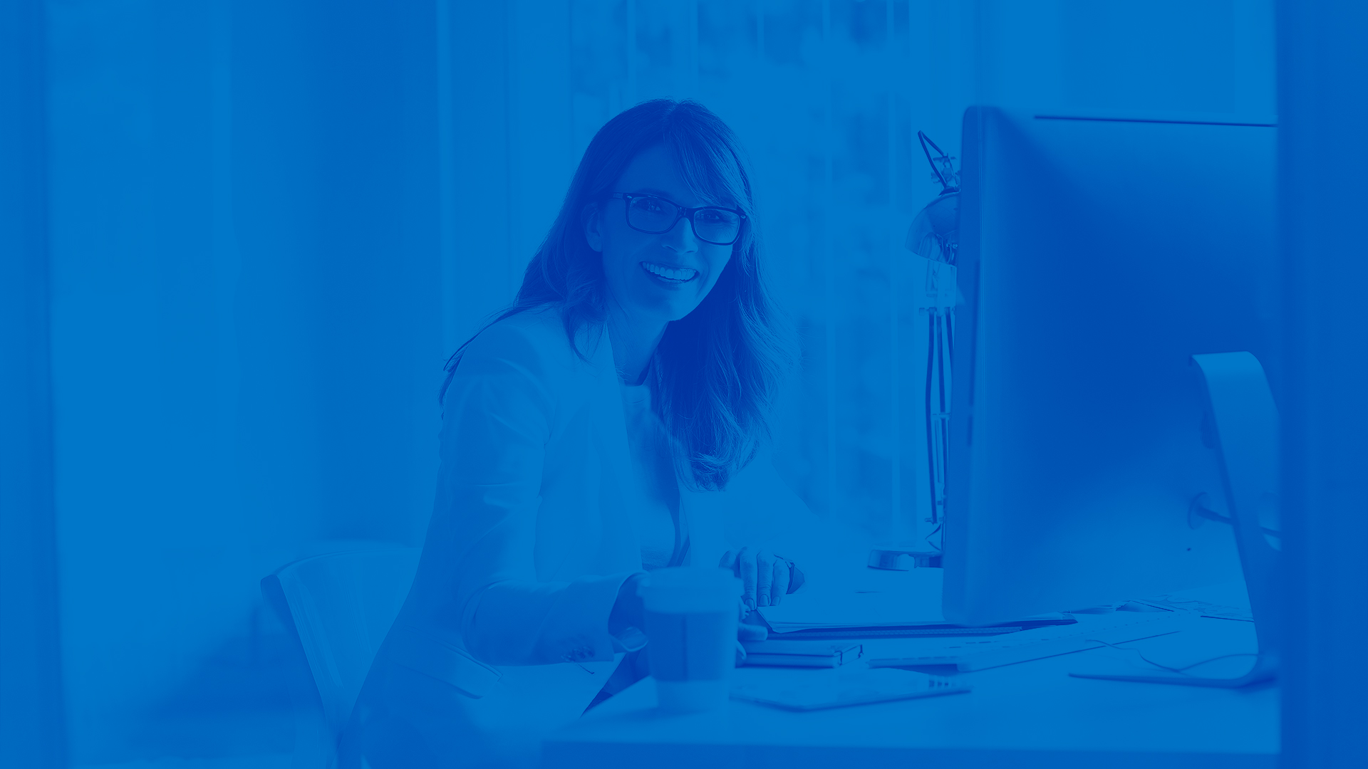 This is an image of a woman sitting at a desk with a desktop computer. She is facing the camera and smiling. There is a blue overlay on the entire image.