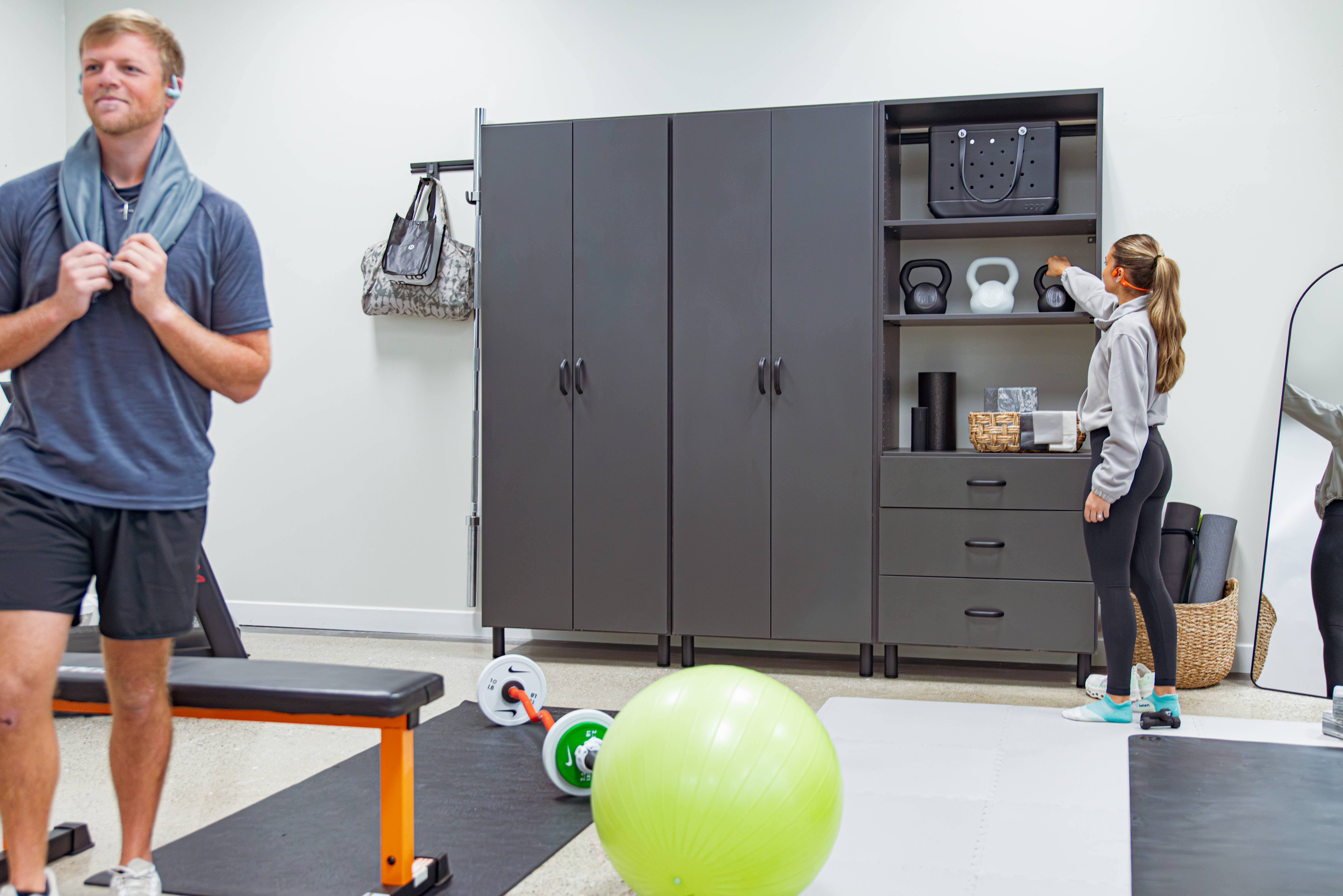Man and woman in a home gym set up with workout bench, exercise ball, weights, and garage system.